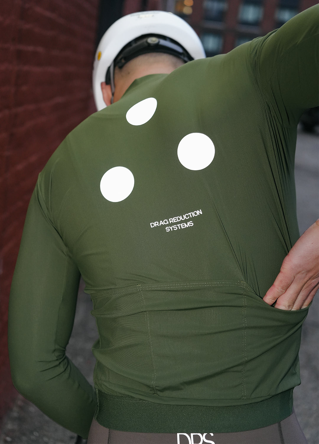 Person wearing a DRS green long-sleeve jersey with white 3-dots and text on the back, standing against a blurred background in Brooklyn, New York. 