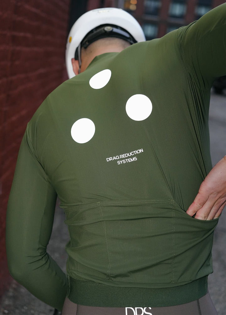 Person wearing a DRS green long-sleeve jersey with white 3-dots and text on the back, standing against a blurred background in Brooklyn, New York. 